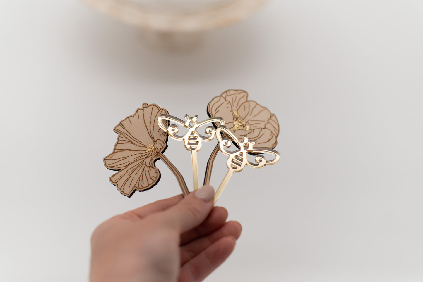 Hand hält 4 Caketopper, 2 goldene Bienen und 2 Holzblumen