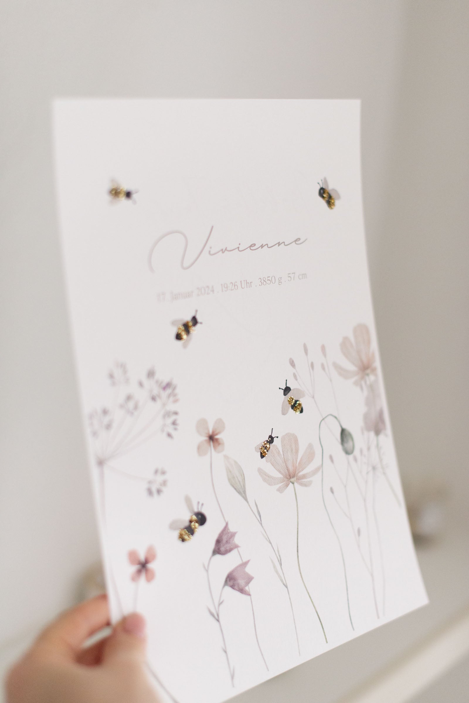 Hand hält Geburtsposter mit glitzernden Bienen und Blumen